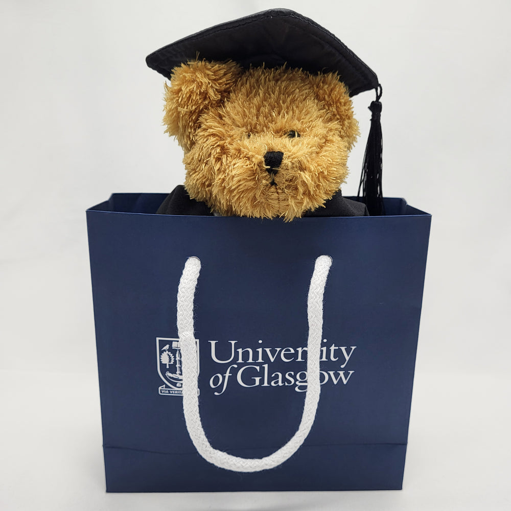University of Glasgow Graduation Bear - Wearing graduation clothing. In UofG navy gift bag, bears face peaks out from top of bag. 