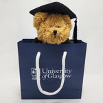 University of Glasgow Graduation Bear - Wearing graduation clothing. In UofG navy gift bag, bears face peaks out from top of bag. 