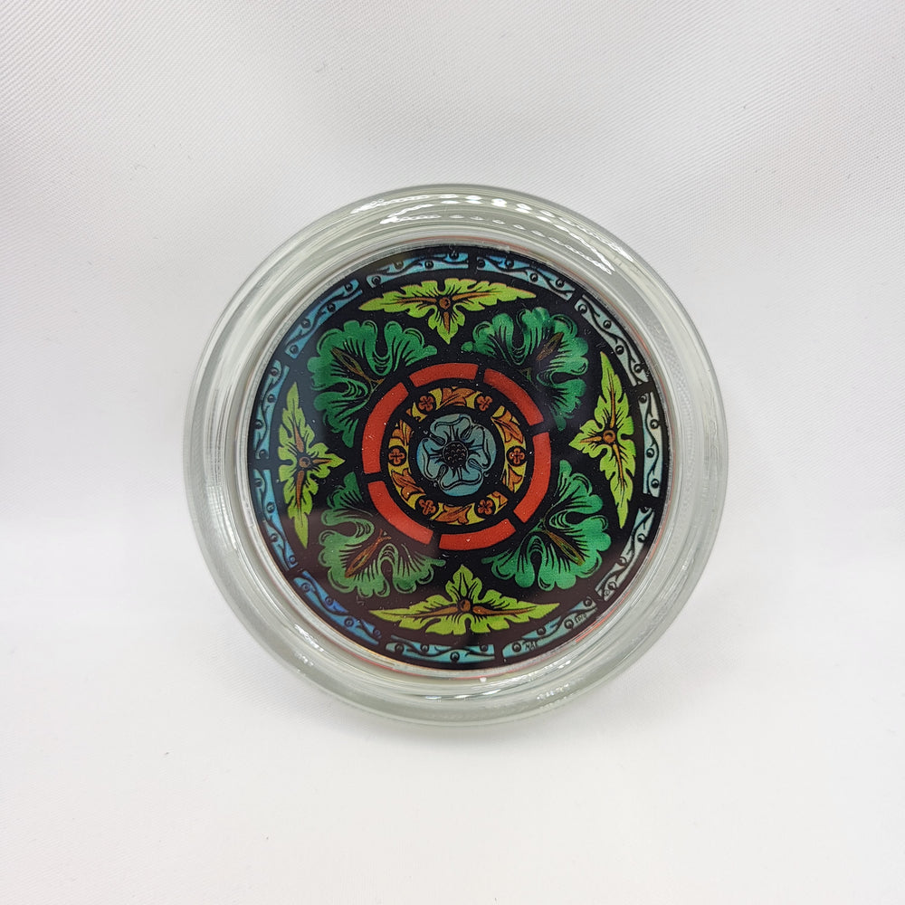 Round glass paperweight. The image shows the paperweight displayed face-on. The paperweight is clear glass with a full-colour stained glass motif in the centre. The motif shows a four-leafed green foliage design with a blue border and blue flower at the centre.