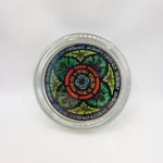 Round glass paperweight. The image shows the paperweight displayed face-on. The paperweight is clear glass with a full-colour stained glass motif in the centre. The motif shows a four-leafed green foliage design with a blue border and blue flower at the centre.
