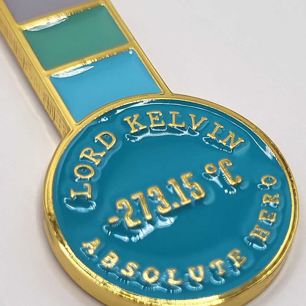 Lord Kelvin Absolute Hero Thermometer Keyring (detail). A full-colour enamel keyring in the shape of a thermometer with a gilt metal chain and key loop. This image shows a close-up of the base of the thermometer, which has raised text in gilt metal against a blue enamel background. The text reads: "Lord Kelvin Absolute Hero" in uppercase letters, with the figure -273.15c in the middle.