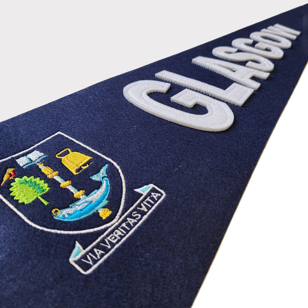 University Crest Pennant Flag (close up view). The image shows a close-up of the full-colour University of Glasgow crest, which is embroidered on the body of the pennant. The word 'GLASGOW' is stitched horizontally next to the crest in white felt. The pennant itself is navy blue felt.