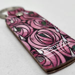 Recycled leather keyring featuring a rose and teardrop design by Charles Rennie Mackintosh. The design features a pattern of pink roses with green accents and white teardrops. The keyring is rectangular with a stainless steel loop at the top. The image shows a close up of the bottom of the keyring, where the Hunterian logo is printed in white.