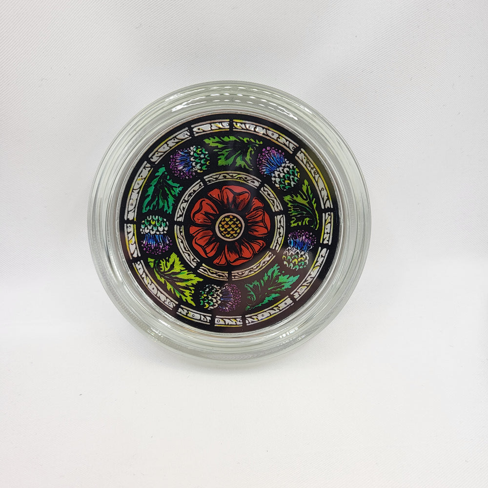 Round glass paperweight. The image shows the paperweight face-on. The paperweight is clear glass with a full-colour stained glass motif in the centre. The motif consists of purple thistles and green leaves in a circular pattern around a single red rose.