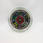 Round glass paperweight. The image shows the paperweight face-on. The paperweight is clear glass with a full-colour stained glass motif in the centre. The motif consists of purple thistles and green leaves in a circular pattern around a single red rose.