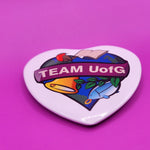 Heart-shaped button badge with 'TEAM UofG' text on a purple background
