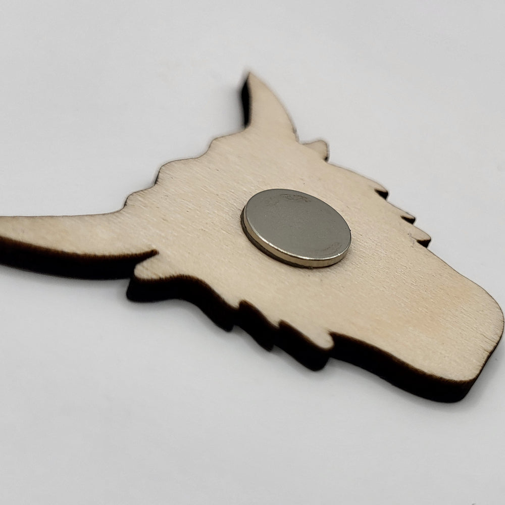Wooden fridge magnet in the shape of a highland cow's head (reverse view). The reverse view shows the circular magnet and the unfinished wooden surface.
