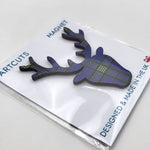 Wooden fridge magnet in packaging. The magnet is in the shape of a stag's head, patterned with the University of Glasgow tartan. The packaging is a white backing card with a clear cellophane wrapping. The card reads: "Artcuts Magnet" along the top and "Designed and made in the UK" along the bottom.