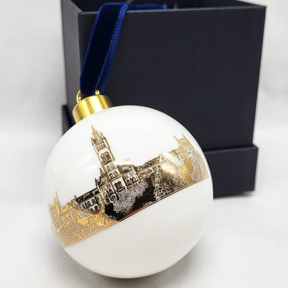 University of Glasgow Tree Bauble - White bauble with gold image of Gilbert Scott Building. In front of black presentation box. Featuring a gold crown attachment and blue velvet ribbon. 