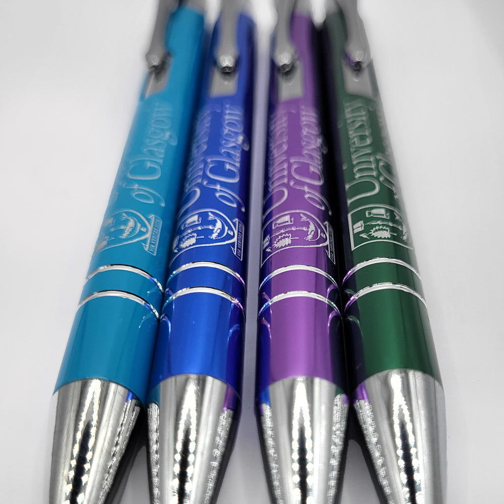 Four metal ball point pens with click tops and a high-gloss finish. From left to right, the pens are: aqua, navy, purple, and green. The UofG crest is printed on the barrel of each pen.