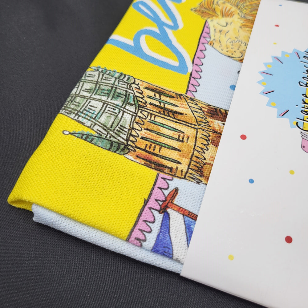 Brightly-coloured tea towel with a Glasgow-themed print, including the Gilbert Scott Building and the tower at the University of Glasgow. The image shows a detail of the teatowel, which is folded up with a cardboard packaging band across the middle.