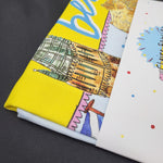 Brightly-coloured tea towel with a Glasgow-themed print, including the Gilbert Scott Building and the tower at the University of Glasgow. The image shows a detail of the teatowel, which is folded up with a cardboard packaging band across the middle.