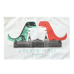 Kelvin Hall Dino Tea Towel. A white cotton tea towel with a colour print in green, red and grey. The illustration shows a t-rex dinosaur facing a t-rex skeleton with the Kelvin Hall building below them. The text below the building reads "Kelvin Hall Glasgow" in block capitals.